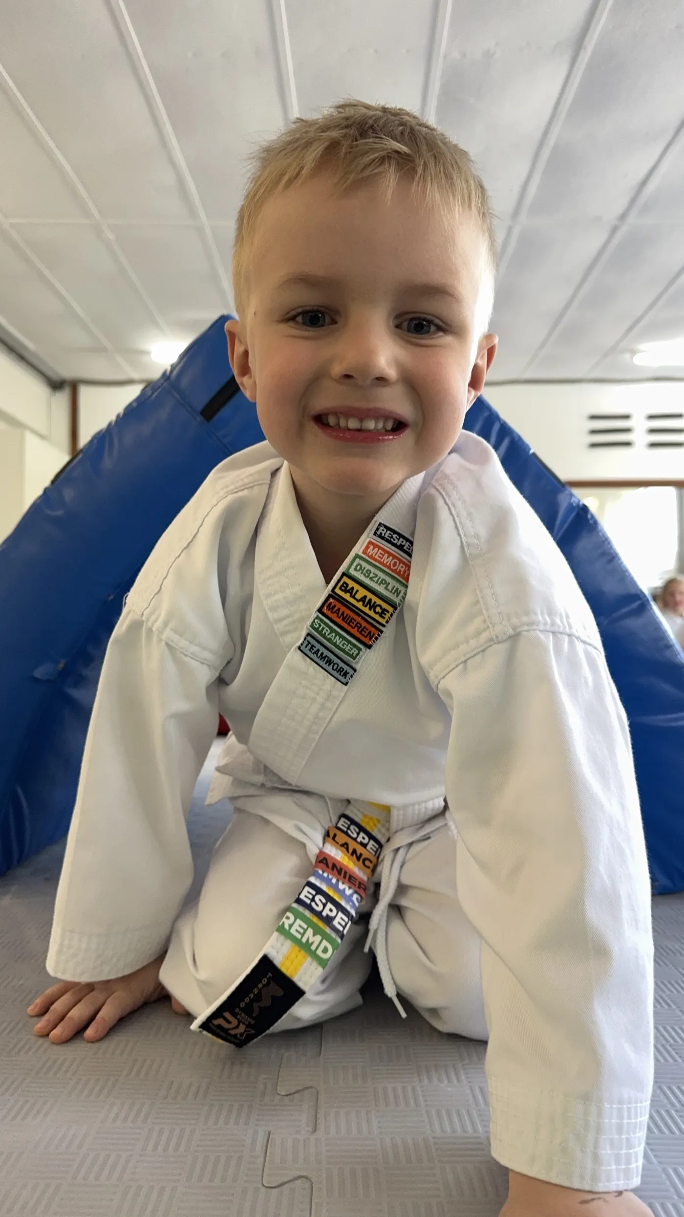 Lachender Junge im weißen Karateanzug mit bunten HEROES-Aufnähern sitzt auf der Matte im Dojo.